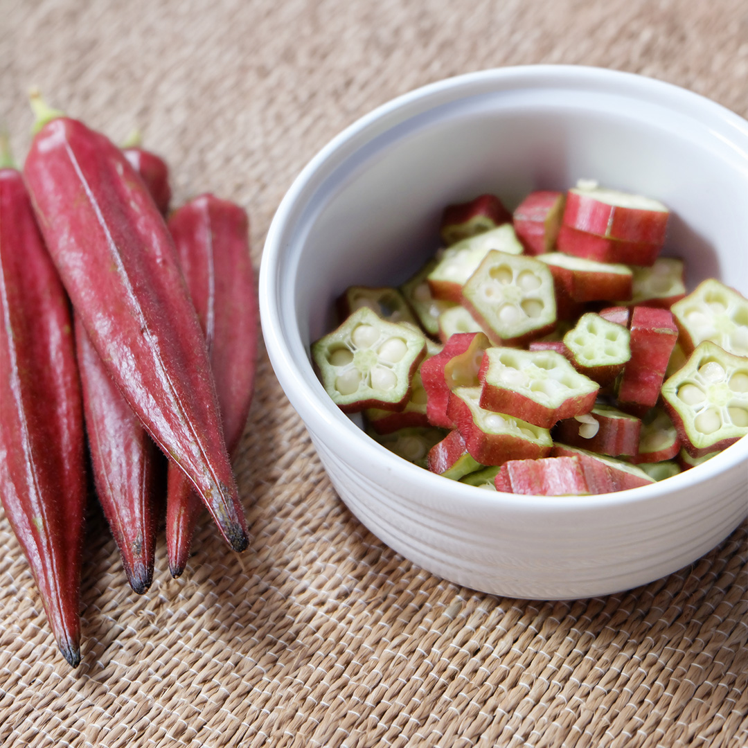 Freshly harvested Burgundy Okra on a woven surface in the kitchen. There is also a white bowl with chopped up red okra in.